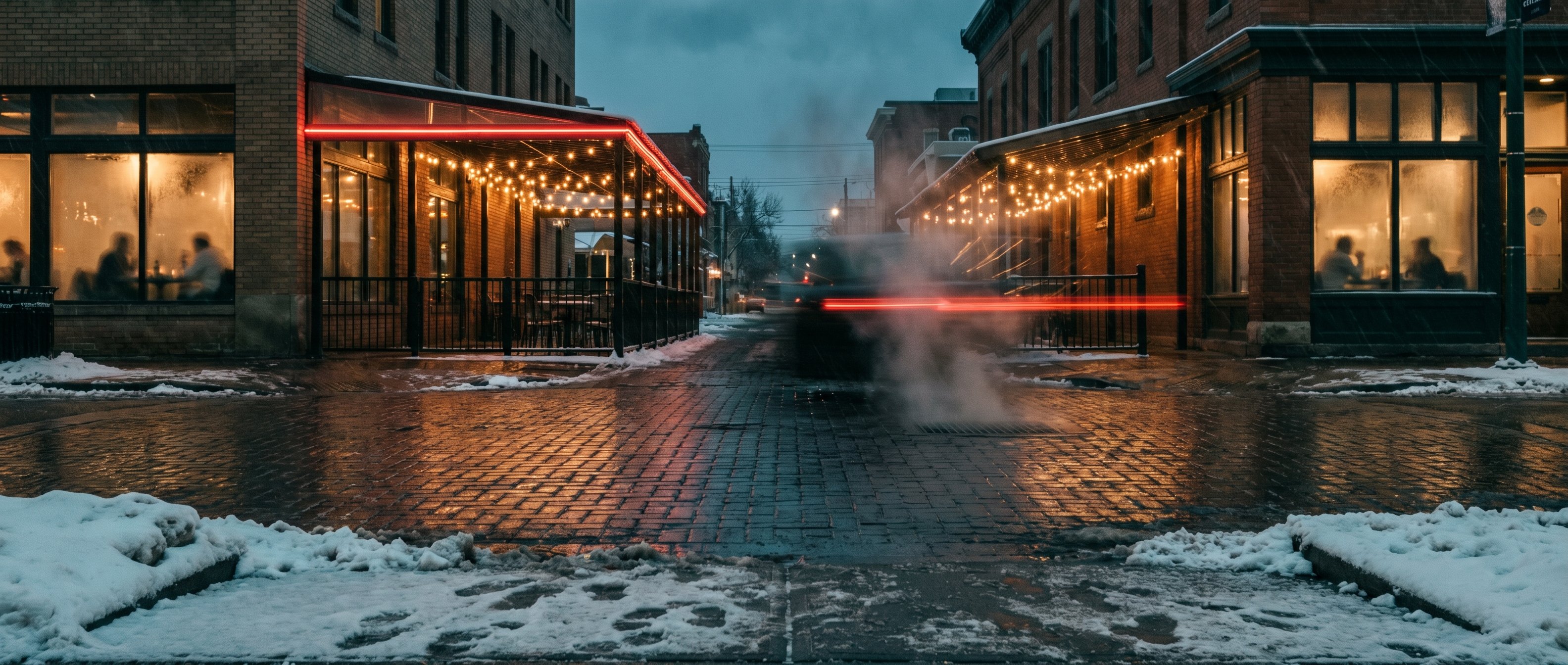 17th Avenue SW Calgary on a Friday evening in winter, wet cobblestone street, restaurant patios on both sides lit with fairy lights, steam rising from a sidewalk grate