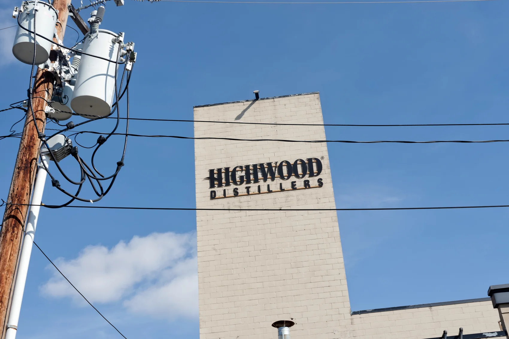 The Highwood Distillers wordmark painted on the white brick tower of the distillery at High River, under a blue sky crossed by power lines. Photograph by Neville Palmer.